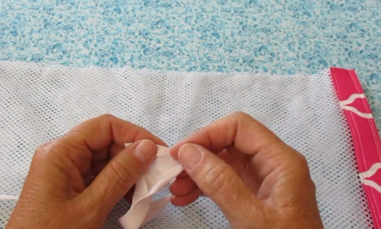A close-up of a sewer's hands folding the end of white bias tape to prepare it for finishing the seam on a mesh laundry bag.