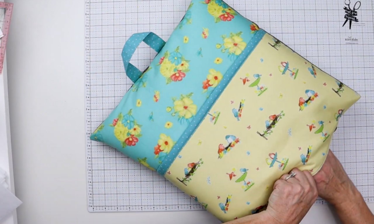 A completed DIY reading pillow with a colorful book pocket made from blue floral and yellow bird-print fabric, sitting on a white gridded cutting mat. A person's hands are stuffing the pillow form into the envelope closure on the back.