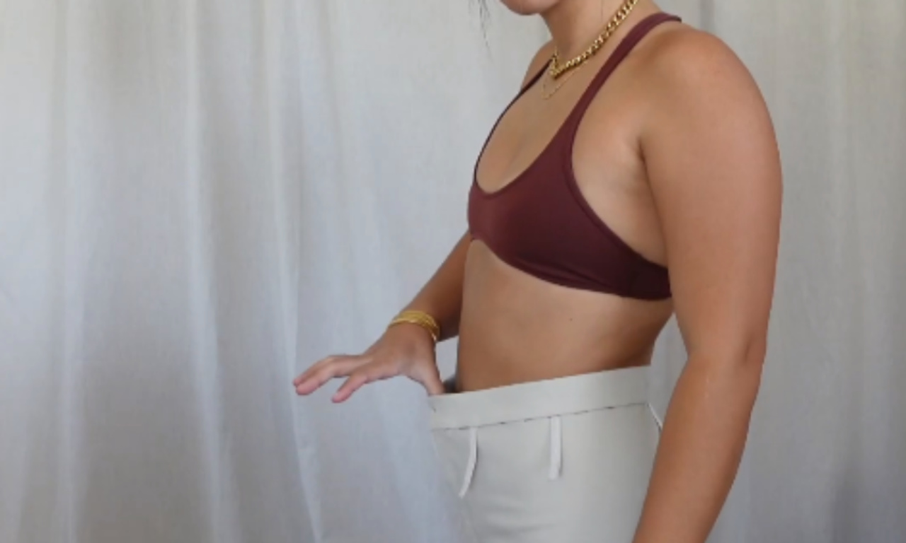 A woman in a brown top pulls the loose waistband of her off-white pants away from her body, showing a large gap and illustrating a common fitting problem.