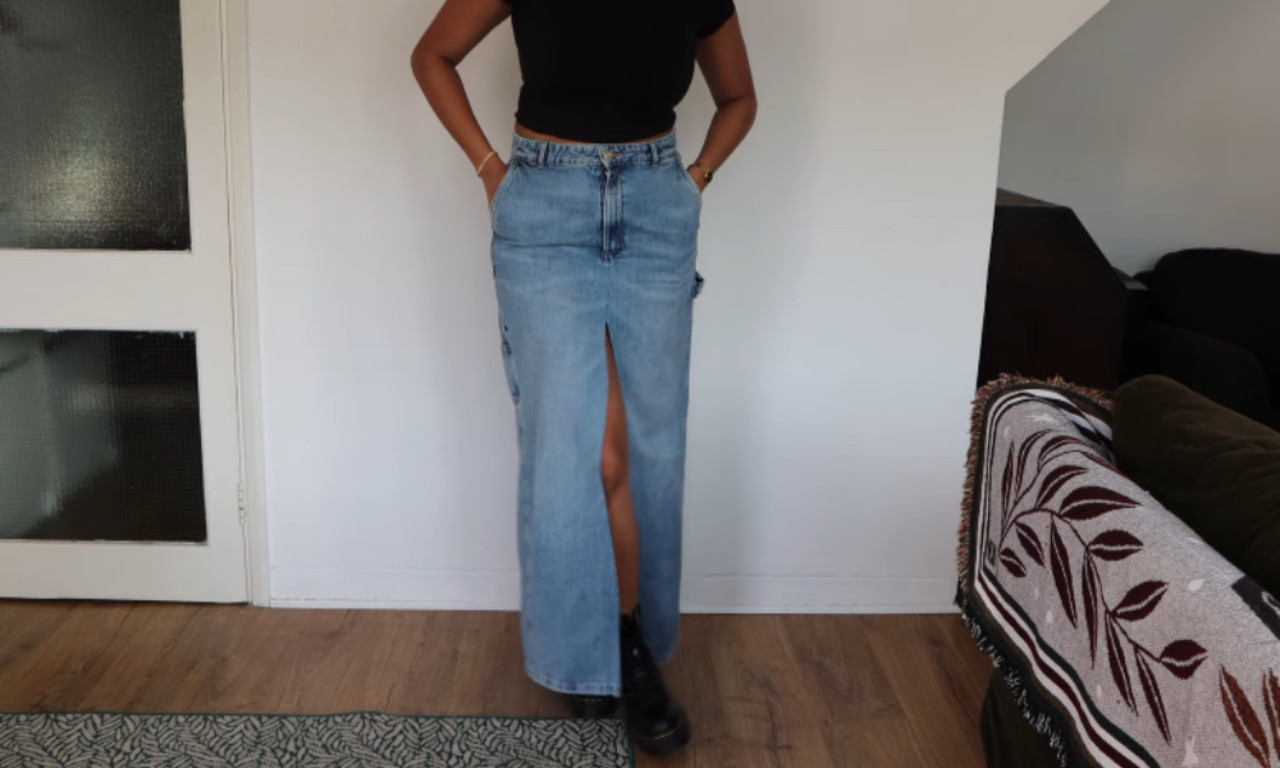 A woman models the final result of a tutorial on how to turn jeans into a skirt, wearing a stylish, long denim maxi skirt with a high front slit and a black top.