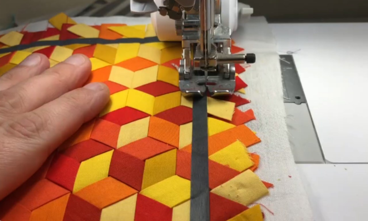 A close-up of a sewing machine stitching a straight line on a piece of fabric with a 3D cube pattern in shades of red, orange, and yellow, with a hand gently guiding the material.