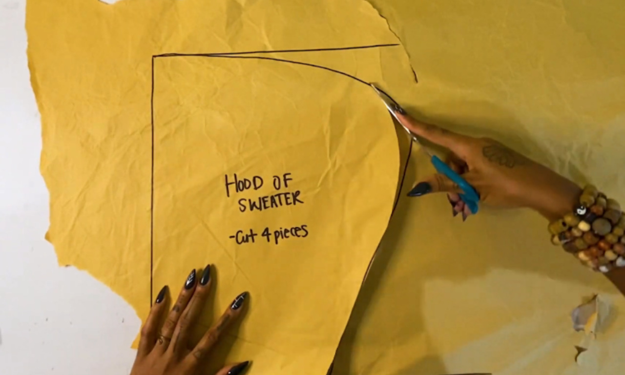 A person cuts a sewing pattern for a hoodie from yellow paper. The pattern piece is labeled "Hood of Sweater - Cut 4 pieces," showing a step in the process of making a DIY hoodie dress from a sweatshirt.