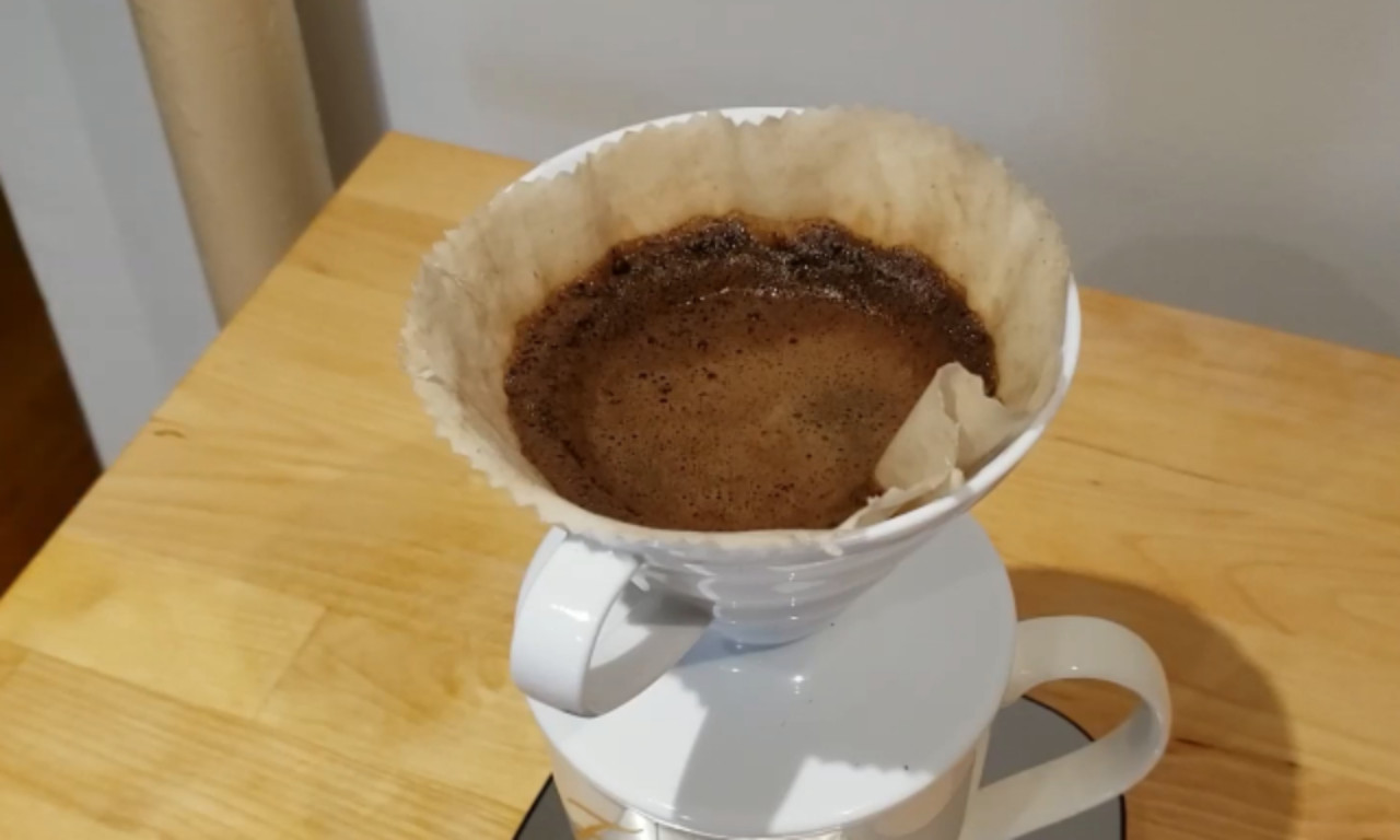 A used reusable fabric coffee filter filled with wet coffee grounds sits inside a white ceramic pour-over dripper on a wooden table.