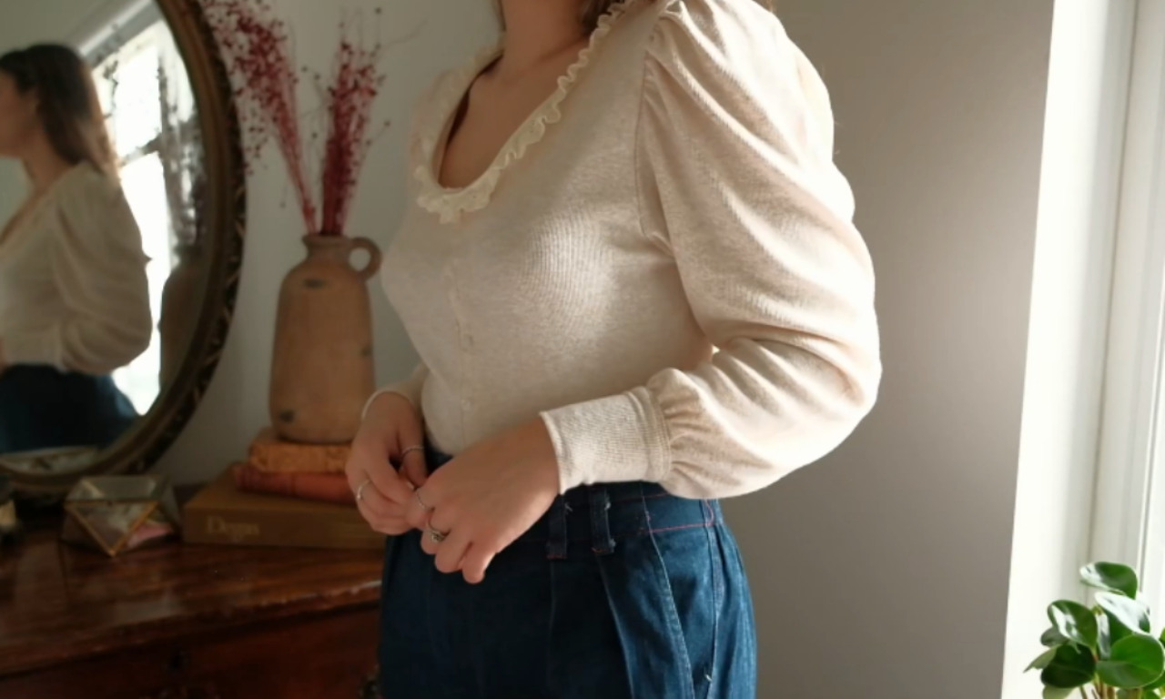 A woman wearing a finished cream-colored knit top with long puff sleeves and a delicate ruffled collar, demonstrating a successful sewing project.