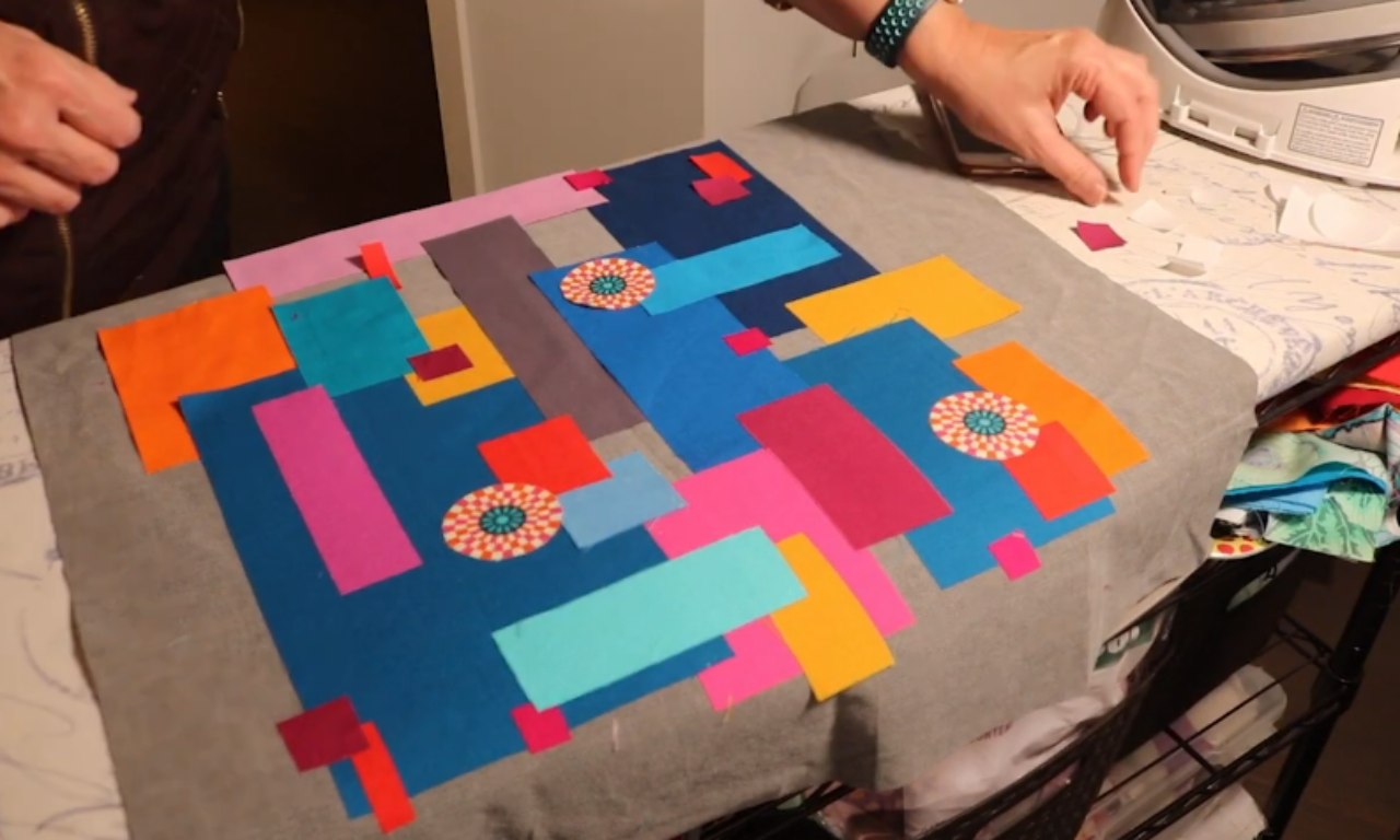 A work in progress of a colorful fabric quilt with irregular geometric shapes like rectangles and squares in bright colors (orange, pink, purple, blue, and teal). Some circular appliqué designs are placed over the patchwork pieces, and an iron and some fabric scraps are visible in the background.