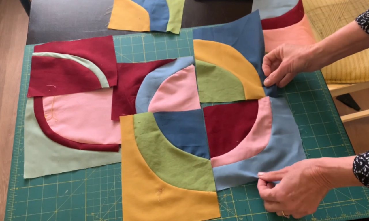 Hands arranging colorful quilt blocks with curved seams on a green cutting mat. The blocks feature fabrics in blue, yellow, green, pink, and burgundy.