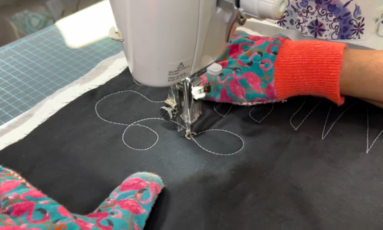 Close-up of a sewing machine stitching white thread in a looping petal design on black fabric, guided by hands wearing colorful quilting gloves.