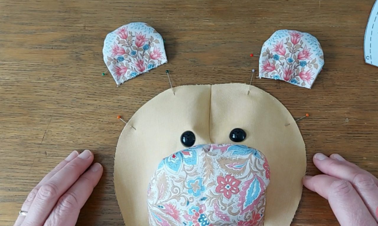 A pair of hands pinning floral fabric ears onto the head of a memory bear during the sewing process.