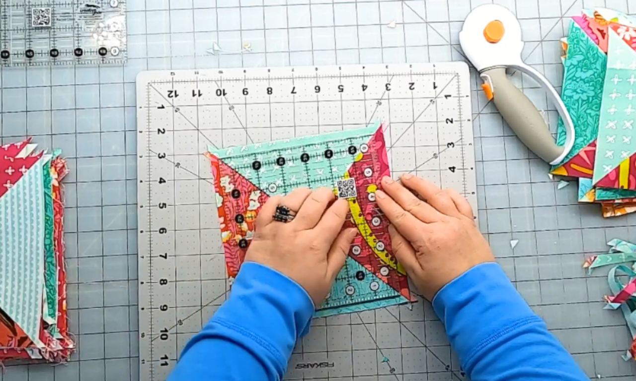 A quilter using a square quilting ruler and a rotary cutter to trim a colorful half-square triangle quilt block on a cutting mat.