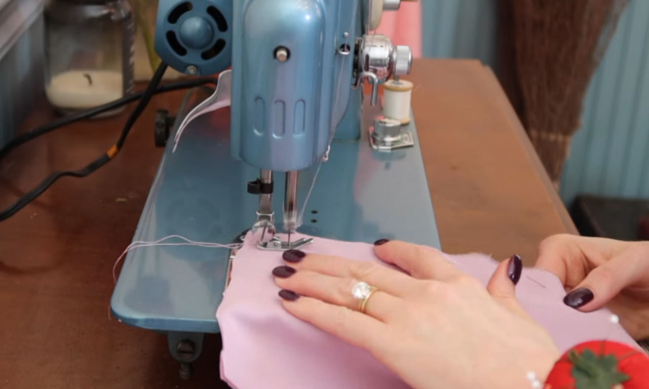 A woman's hands guiding pink satin fabric through a vintage blue sewing machine, demonstrating a key step in sewing vintage coats.