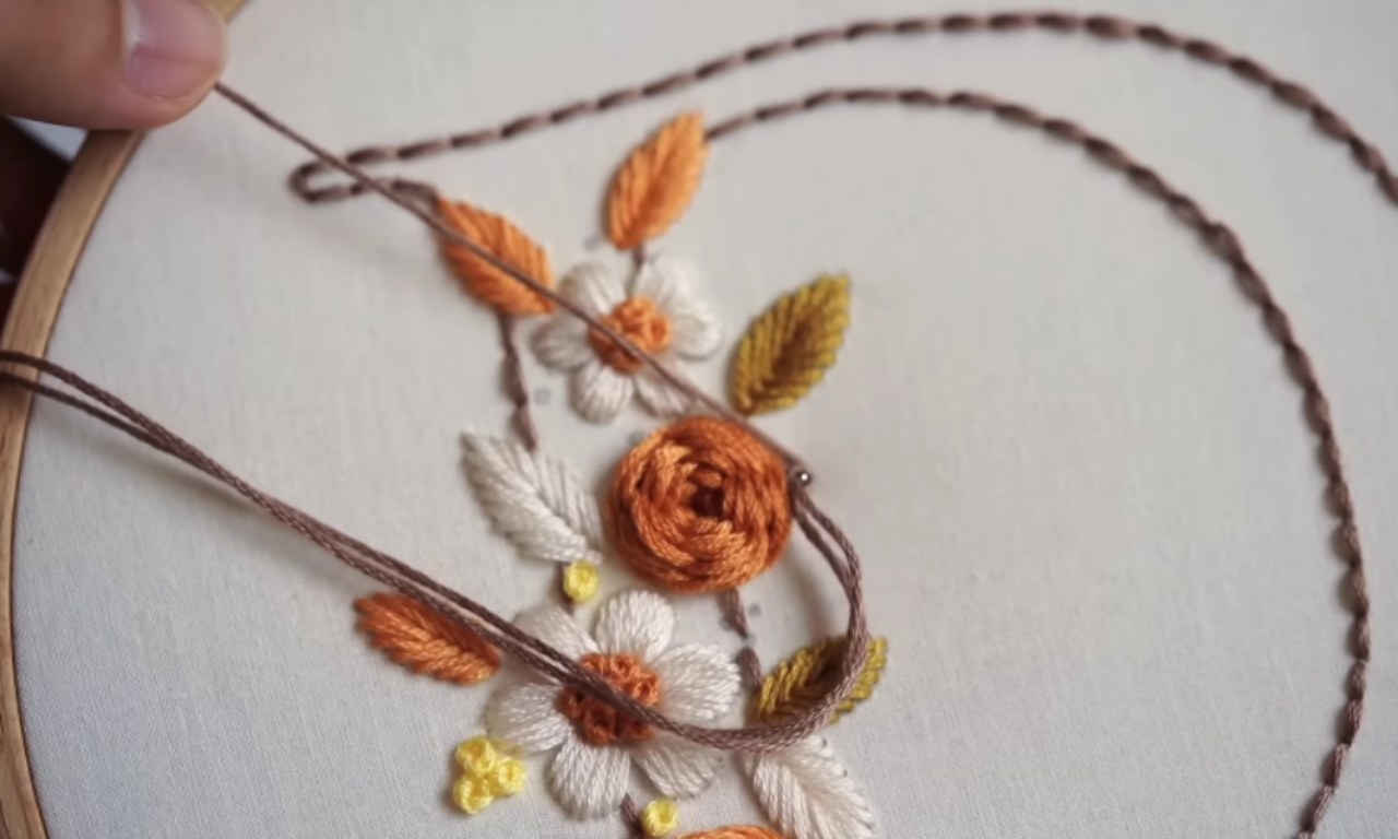 A close-up view of a hand stitching intricate orange and white flowers onto fabric in an embroidery hoop, showing the embroidery process in action.
