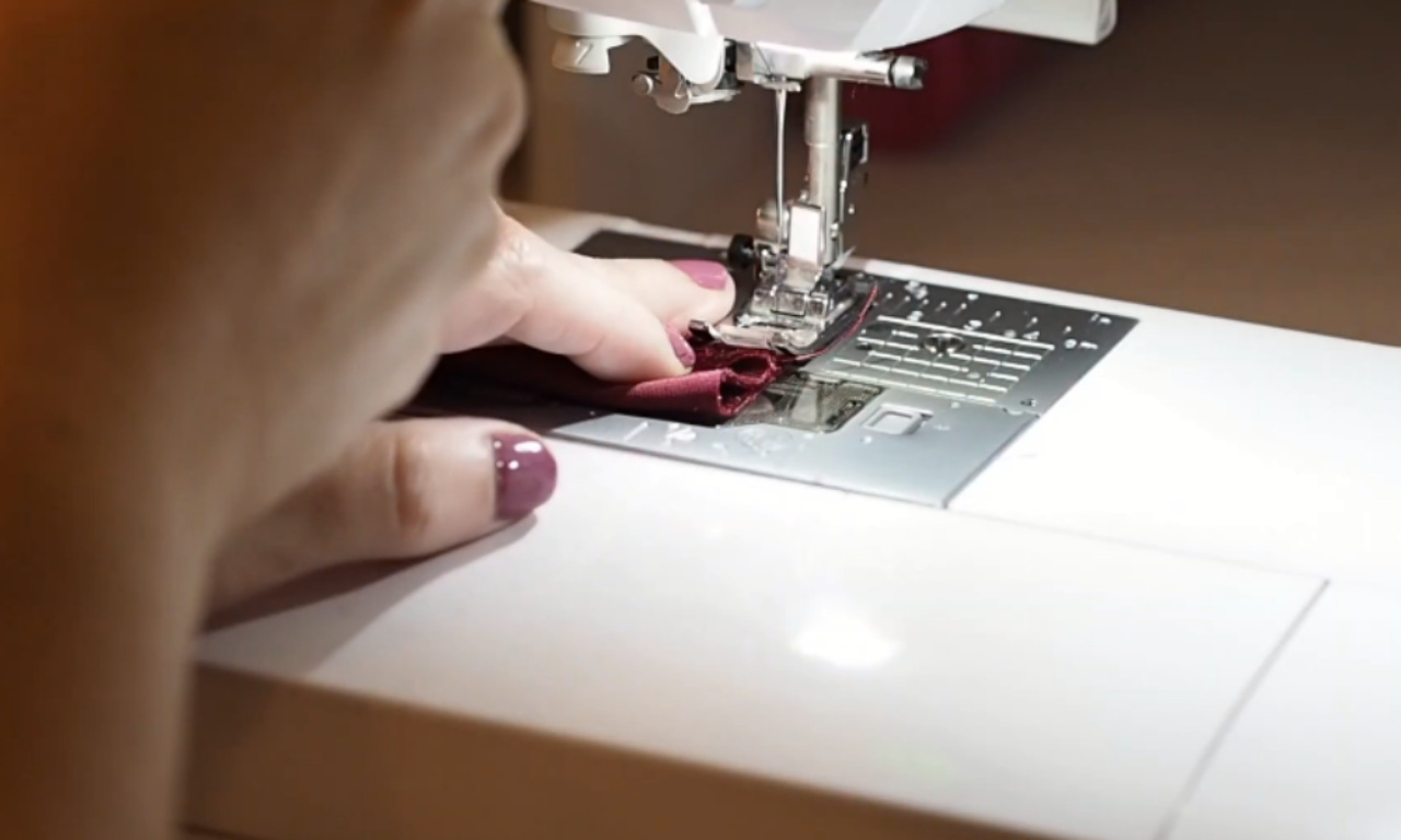 A close-up view of a person sewing burgundy fabric on a white sewing machine, guiding the cloth with their fingers.