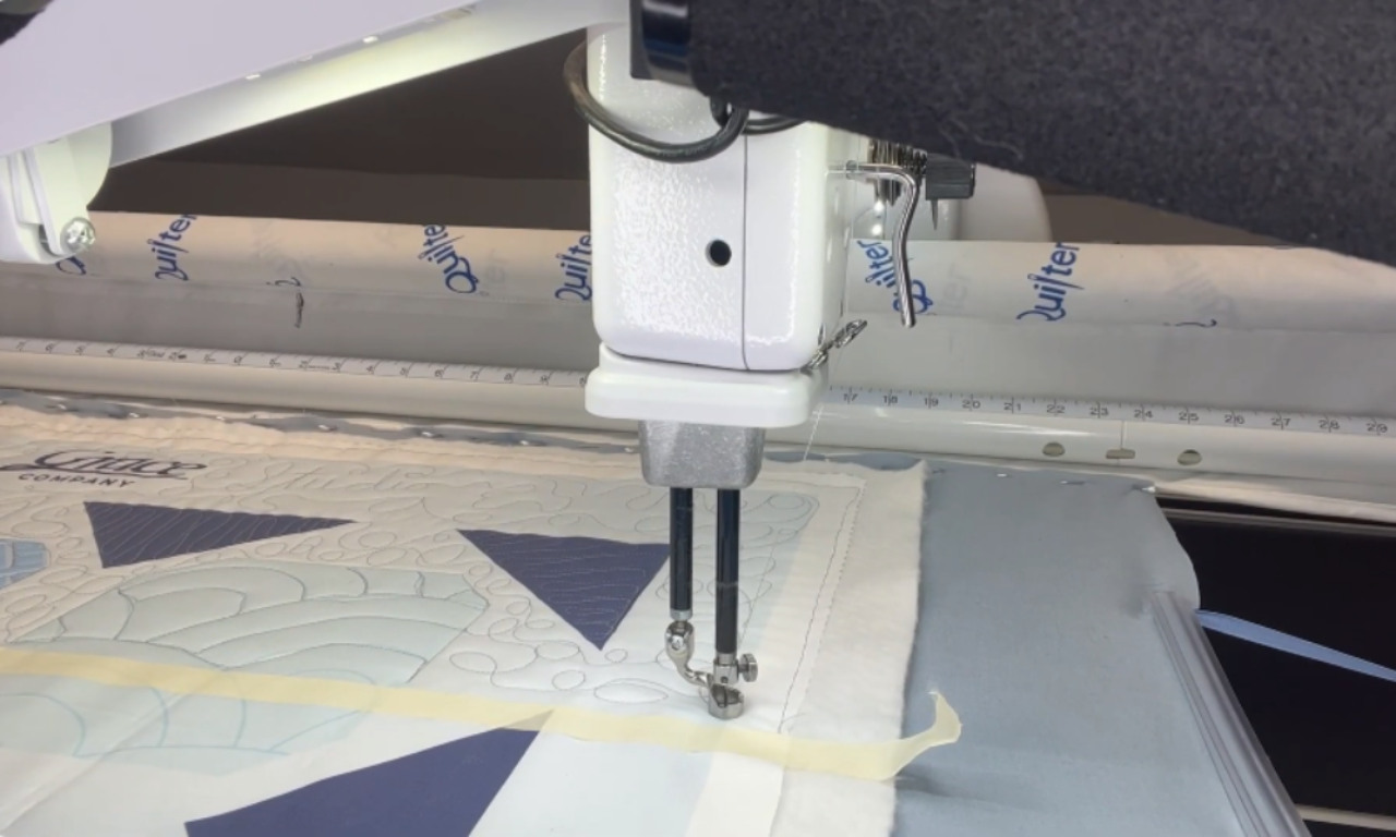 A white longarm quilting machine in action, stitching a decorative pattern onto a blue and white pre-printed quilt panel loaded on a quilting frame.