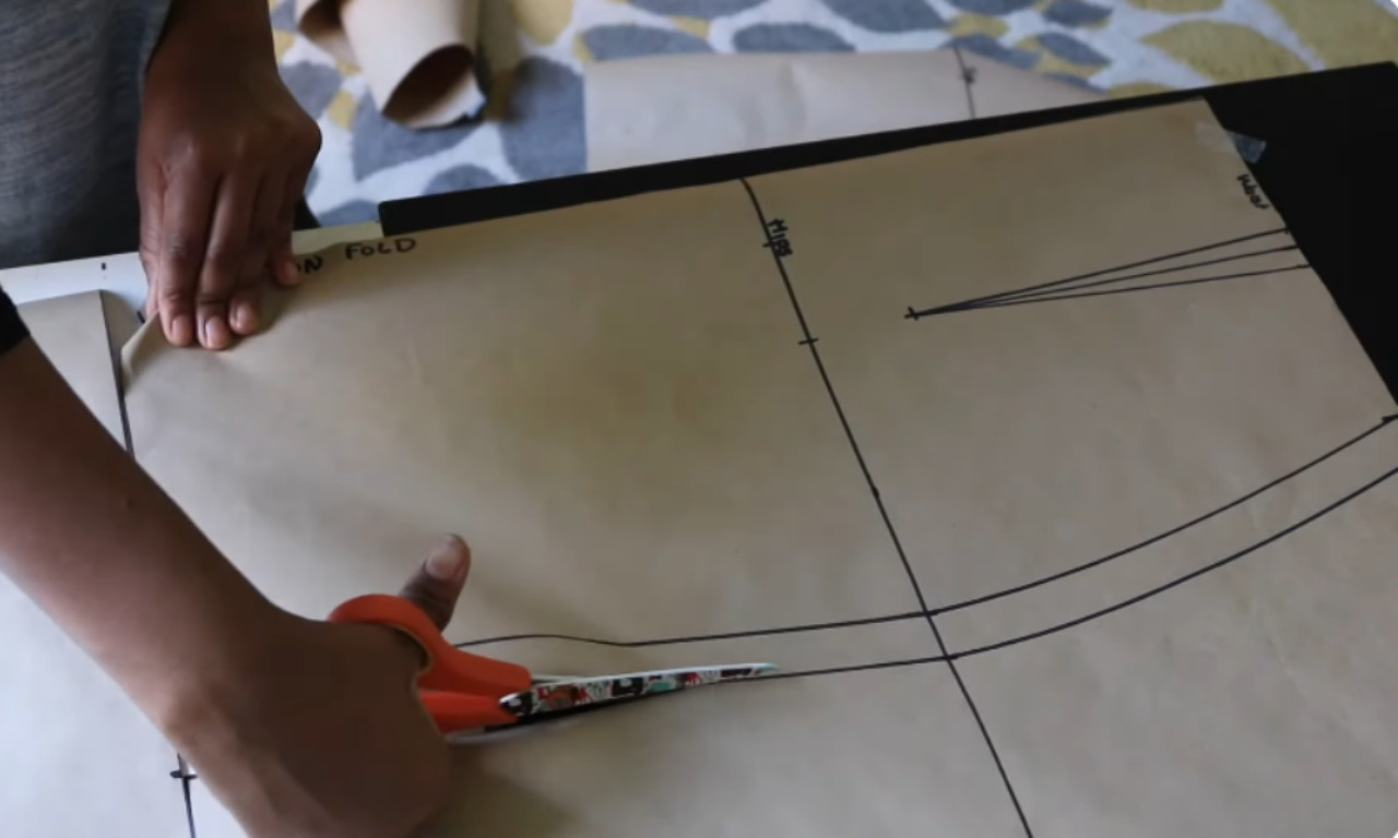 A pair of hands cutting along the drawn outline of a brown paper skirt pattern with a pair of orange-handled scissors.