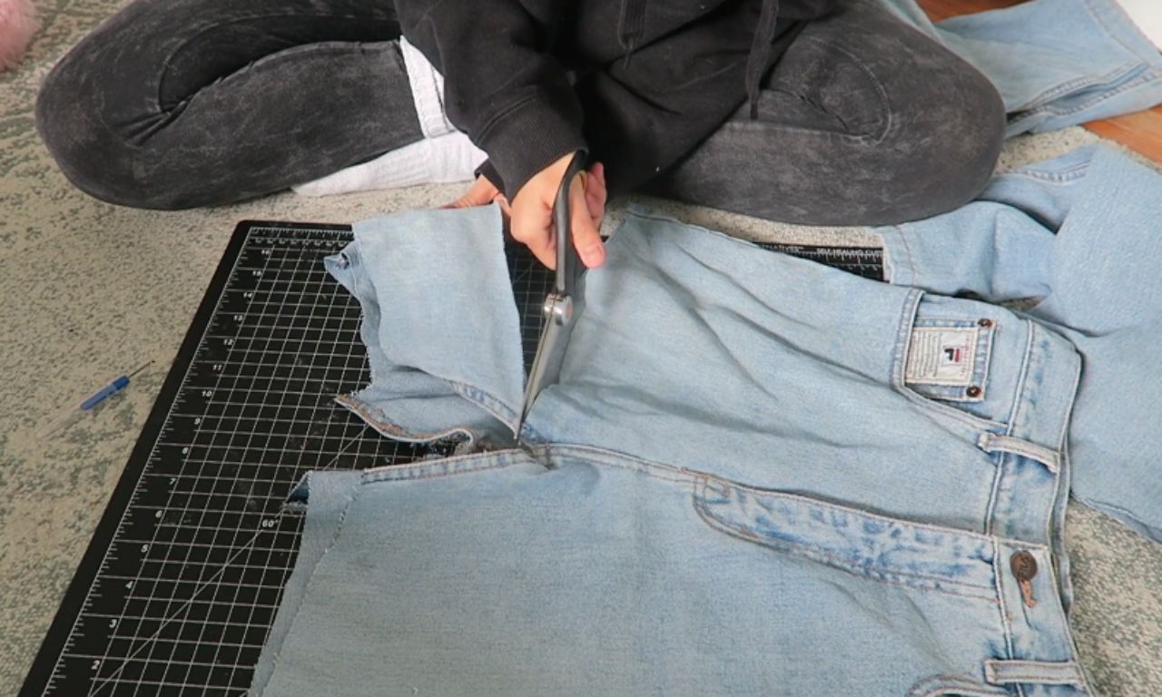 A person sitting on the floor uses sharp fabric scissors to cut the inseam of a pair of light-wash jeans laid on a black cutting mat, demonstrating a key step in how to turn jeans into a skirt.