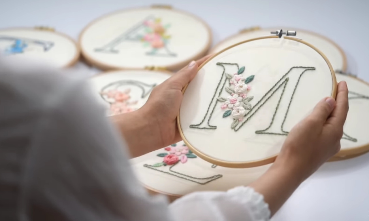 Hands holding up a completed embroidery hoop showcasing the letter M hand-stitched with green thread and adorned with pink and white flowers.