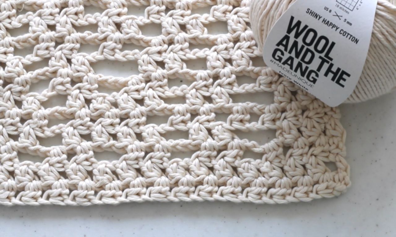 A close-up swatch of the lacy crochet knot stitch pattern in ivory white cotton, with the ball of Wool and the Gang yarn used for the project next to it.