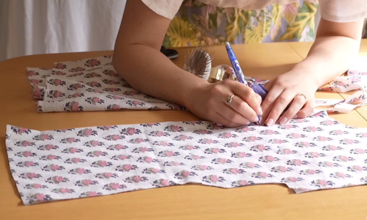 Person using a fabric marker to draw on floral fabric laid out on a table. The fabric features a repeating pattern of pink and purple flowers.