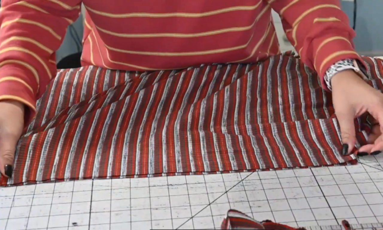 A person in a red striped shirt spreads out a piece of red and white striped fabric on a cutting mat, preparing it for sewing.