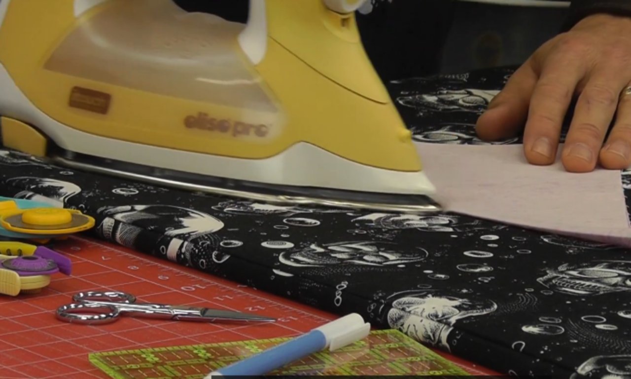 A person using a yellow Oliso Pro iron to press a crease into a quilt block on an ironing board, with sewing tools in the foreground.