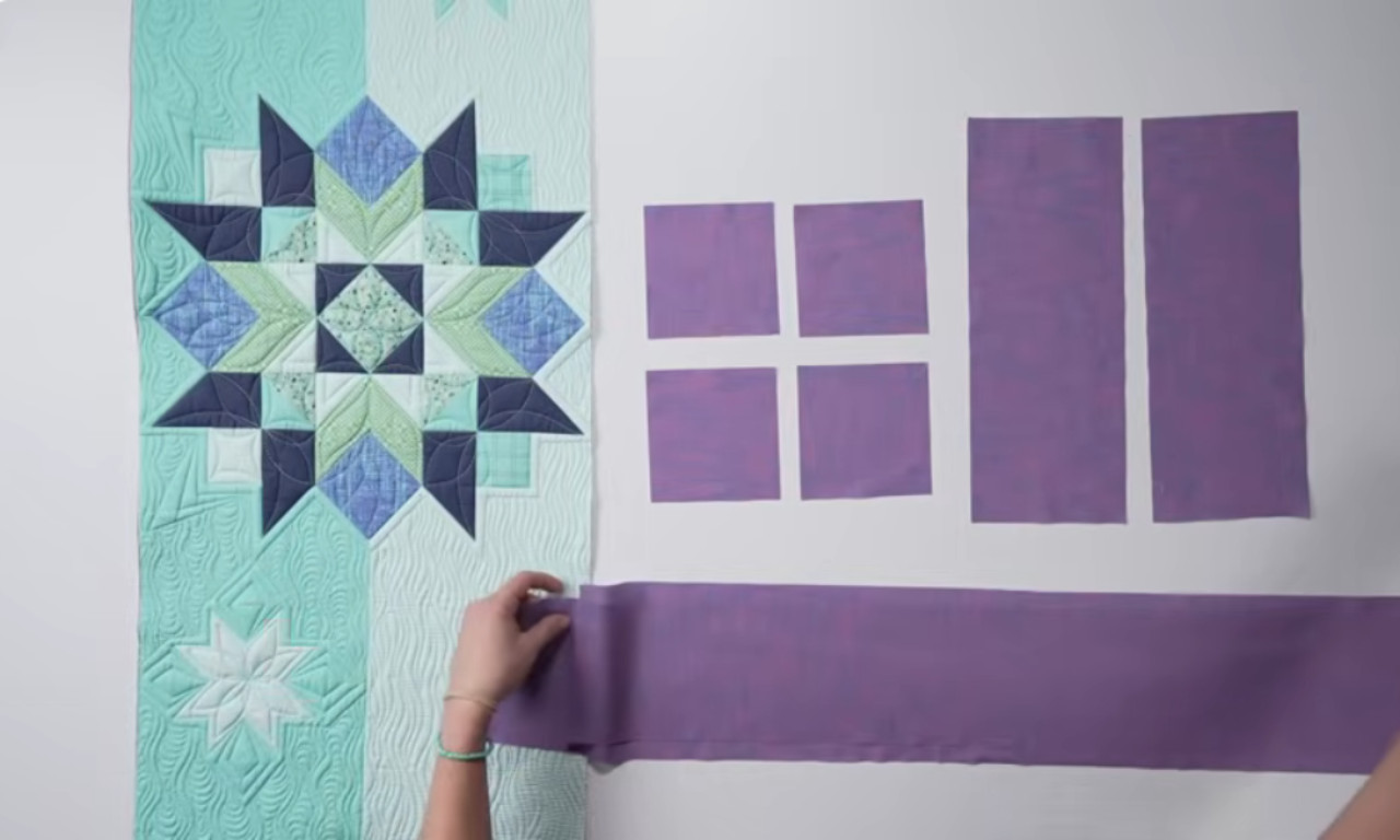 A person arranging purple fabric pieces of different shapes and sizes next to a teal quilt block with a star pattern.
