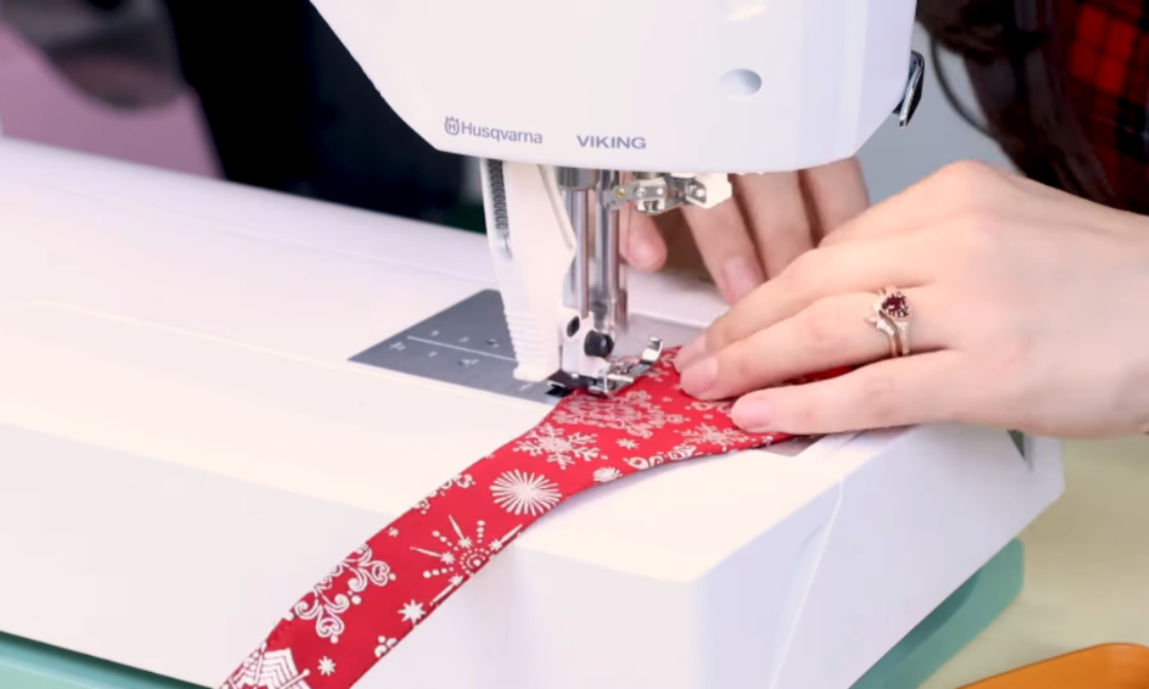 A person's hands guiding a red fabric with a white snowflake pattern under the needle of a white sewing machine to sew a DIY bow tie.