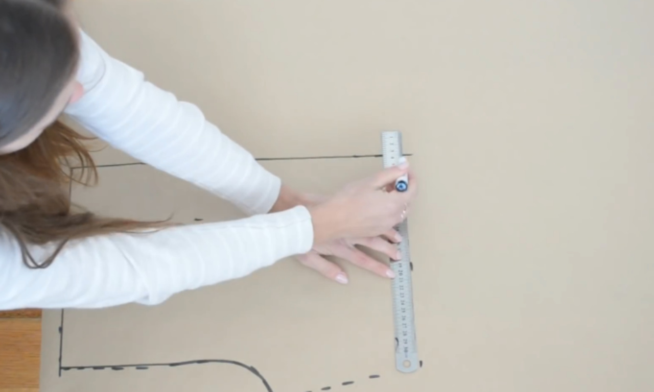 A sewer uses a ruler and a marker to draft a sewing pattern for shorts on a large sheet of brown paper.