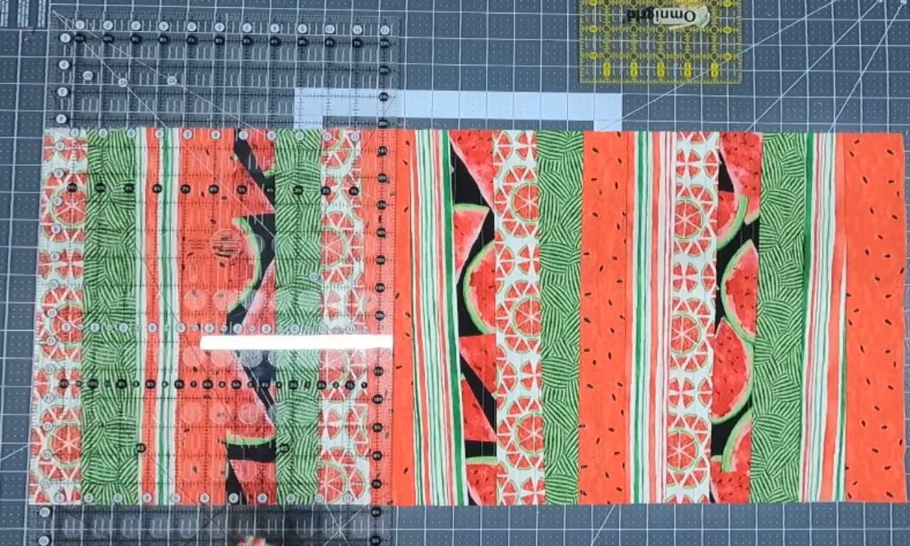 Strips of fabric with watermelon-themed prints laid side by side on a cutting mat, with quilting rulers placed on top, ready for precise cutting.