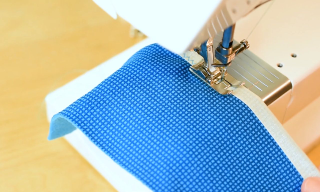 A close-up of a sewing machine stitching a flat, unfolded piece of white fold-over elastic onto the edge of a blue patterned fabric.
