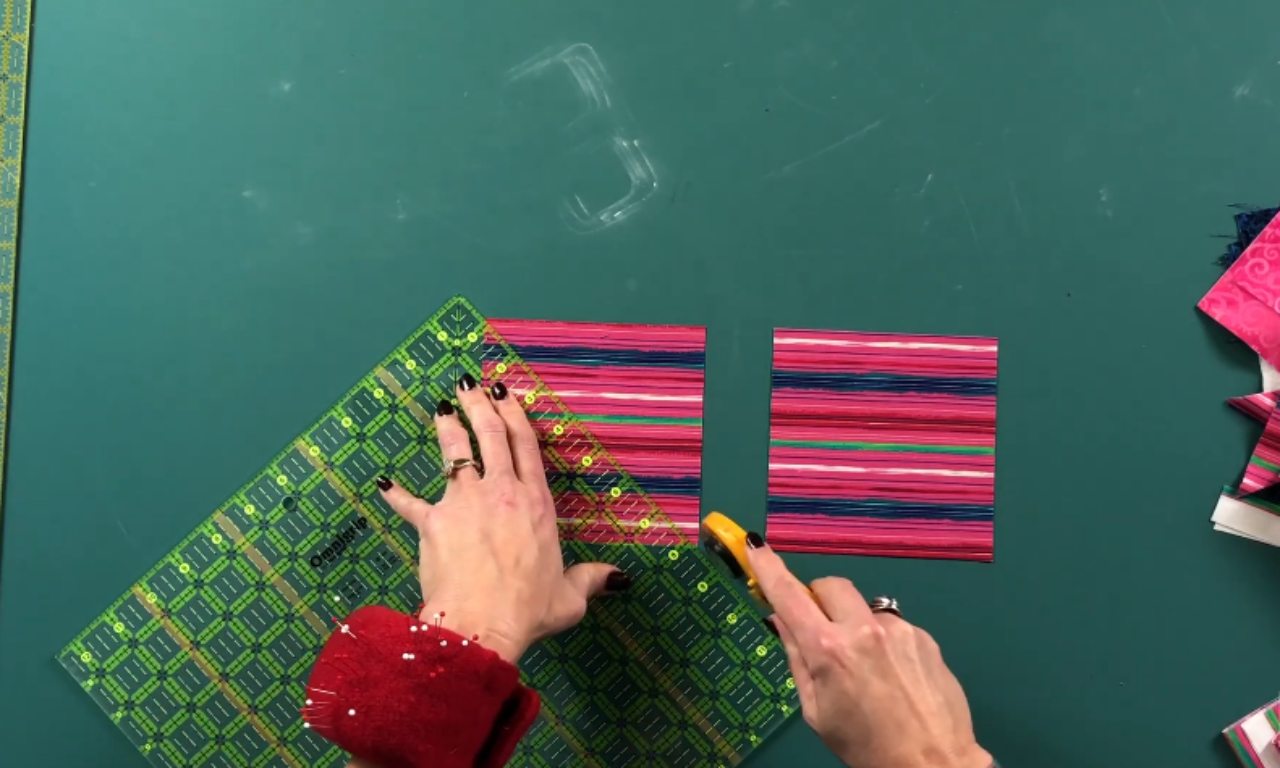 A quilter uses a rotary cutter and a clear quilting ruler to cut a square of pink striped fabric on a green cutting mat.
