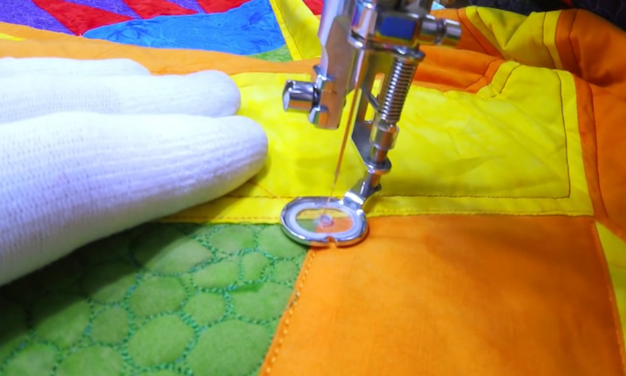 Close-up of a sewing machine doing free-motion quilting on a yellow and orange quilt block, with a gloved hand guiding the fabric.