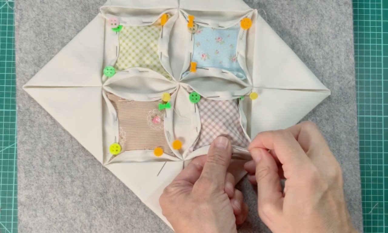 A person hand-sewing a Cathedral Window quilt block, using a needle and thread to stitch the folded white fabric over a pastel-colored scrap square.