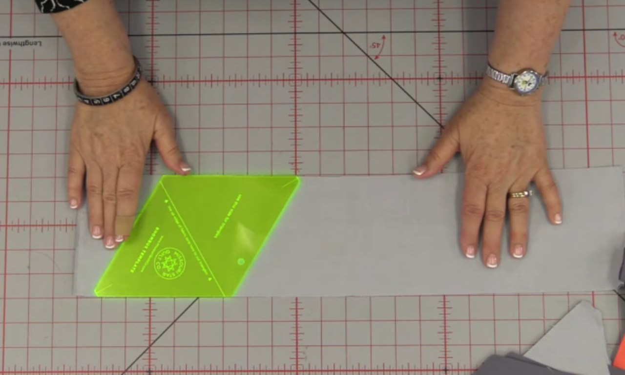 A person is positioning a neon green triangle ruler on a gray fabric strip, preparing to cut precise triangular pieces for a quilt. The cutting mat underneath is marked with red grid lines and angle guides.
