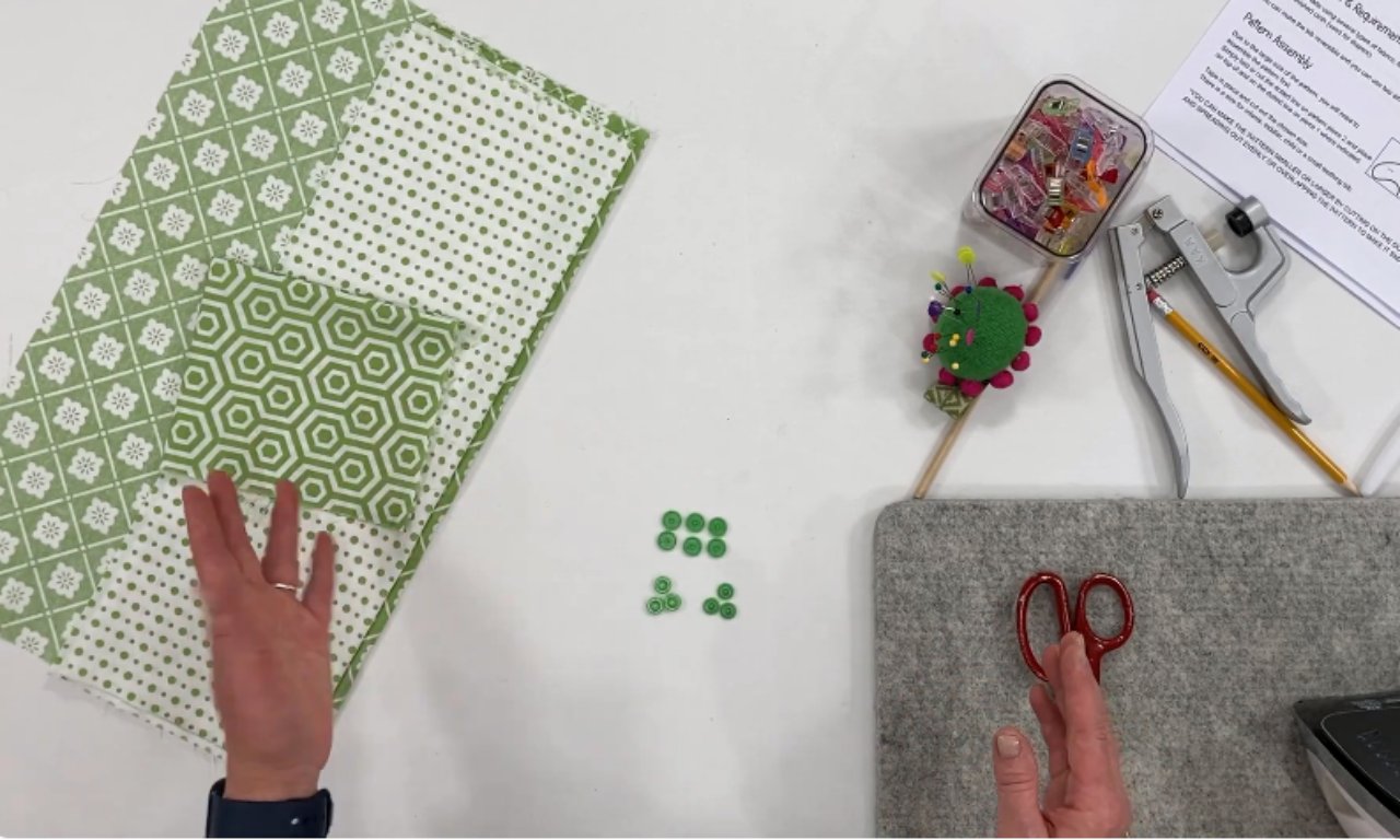 Several pieces of green and white patterned fabric laid out on a table with sewing supplies, including green snap buttons, scissors, a pincushion, sewing clips, and a snap fastener tool. A hand is pointing toward the fabric.