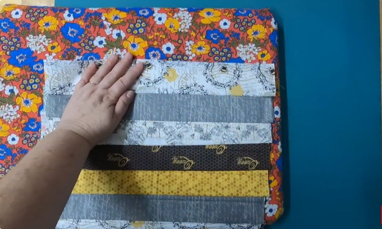 A hand resting on a stack of colorful fabric strips arranged horizontally, featuring floral and patterned designs in shades of gray, white, yellow, and black, placed over a vibrant floral background fabric.