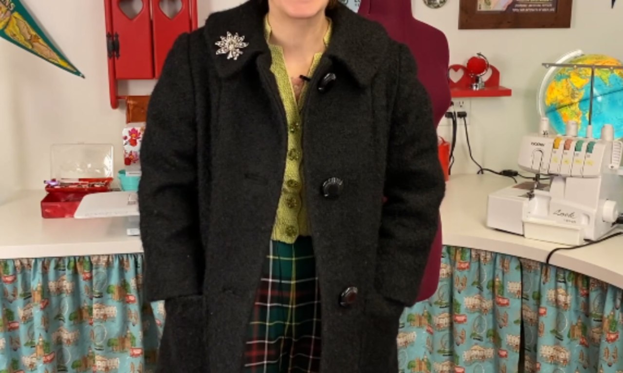 A woman models a thick, black vintage-style wool coat with a Peter Pan collar and large buttons in her sewing studio.