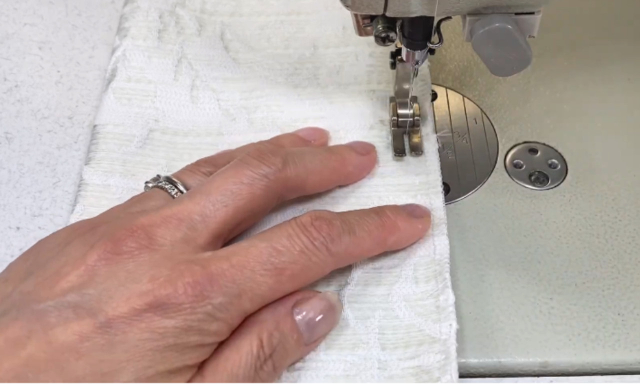 A close-up of a hand sewing white textured fabric on a sewing machine, stitching along the edge.
