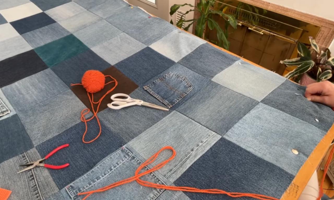 A denim quilt on a wooden frame with tools for quilt tying laid on top, including a ball of orange yarn, scissors, and pliers.