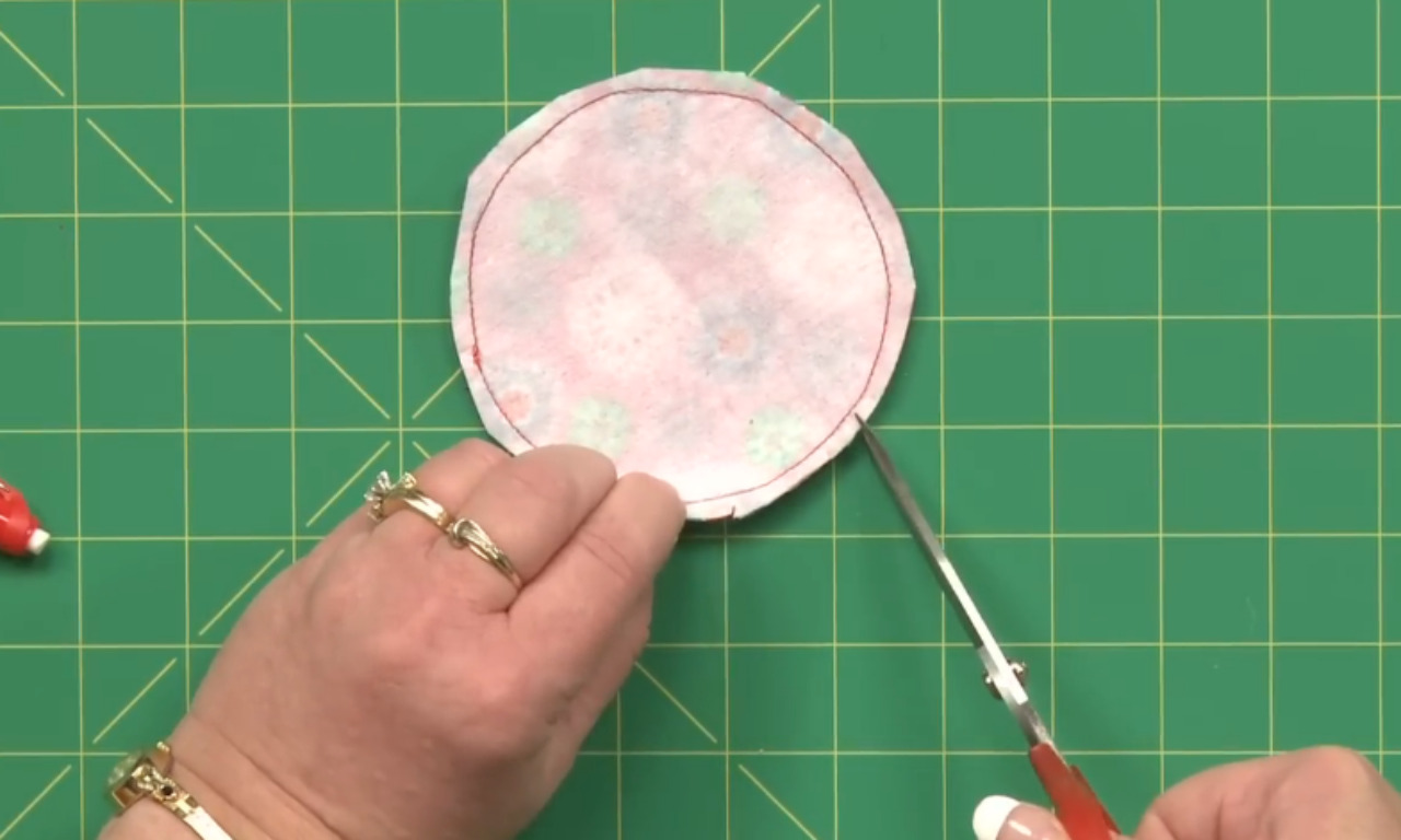 A sewer's hand holds a circular piece of fabric with red stitching around the edge while using a small pair of scissors to clip the curved seam allowance on a green cutting mat.