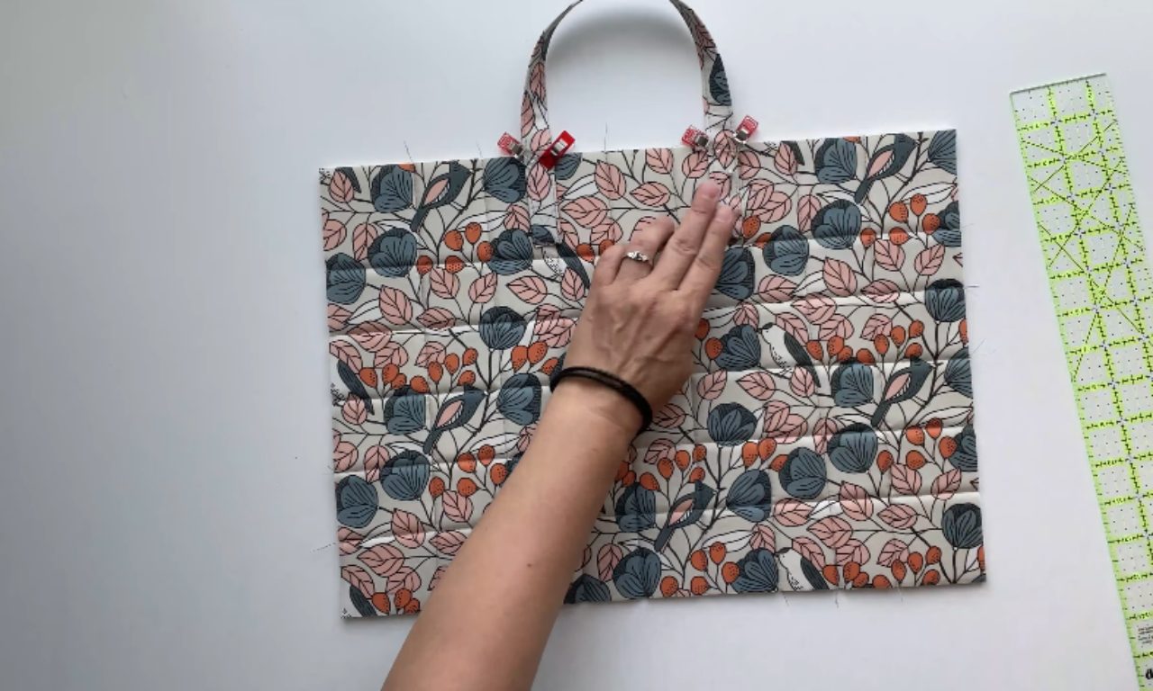A hand smoothing a piece of quilted floral fabric for a laptop bag, with two handles attached at the top with red sewing clips.