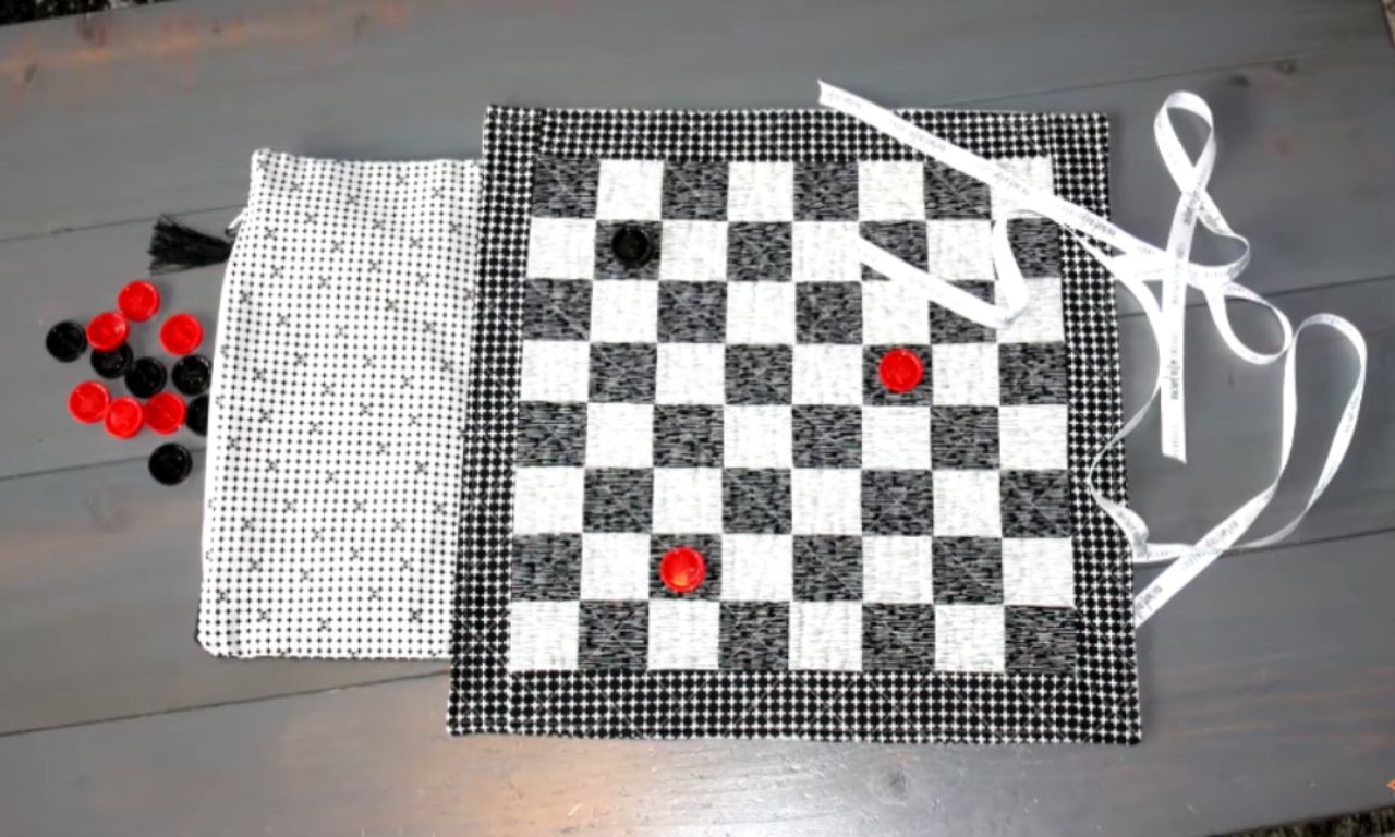 A black-and-white quilted checkerboard laid flat with a few red and black checkers pieces placed on it, next to a matching fabric bag holding the rest of the game pieces. White ribbon ties are attached to the board’s edges.