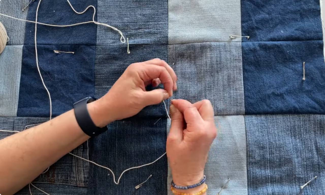 Close-up of a person's hands tying a knot with white yarn on a denim quilt that is held together with safety pins, demonstrating how to tie a quilt.