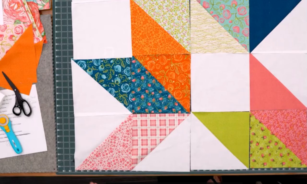 A set of colorful half-square triangle quilt blocks laid out on a cutting mat. The fabrics include floral, plaid, and solid prints in shades of blue, orange, pink, green, and white. Scissors, fabric pieces, and a rotary cutter are placed nearby on the table.
