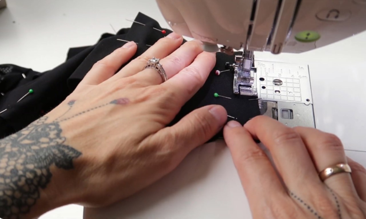 A close-up of hands guiding pinned black fabric panels through a sewing machine to create the seams for DIY mesh leggings.