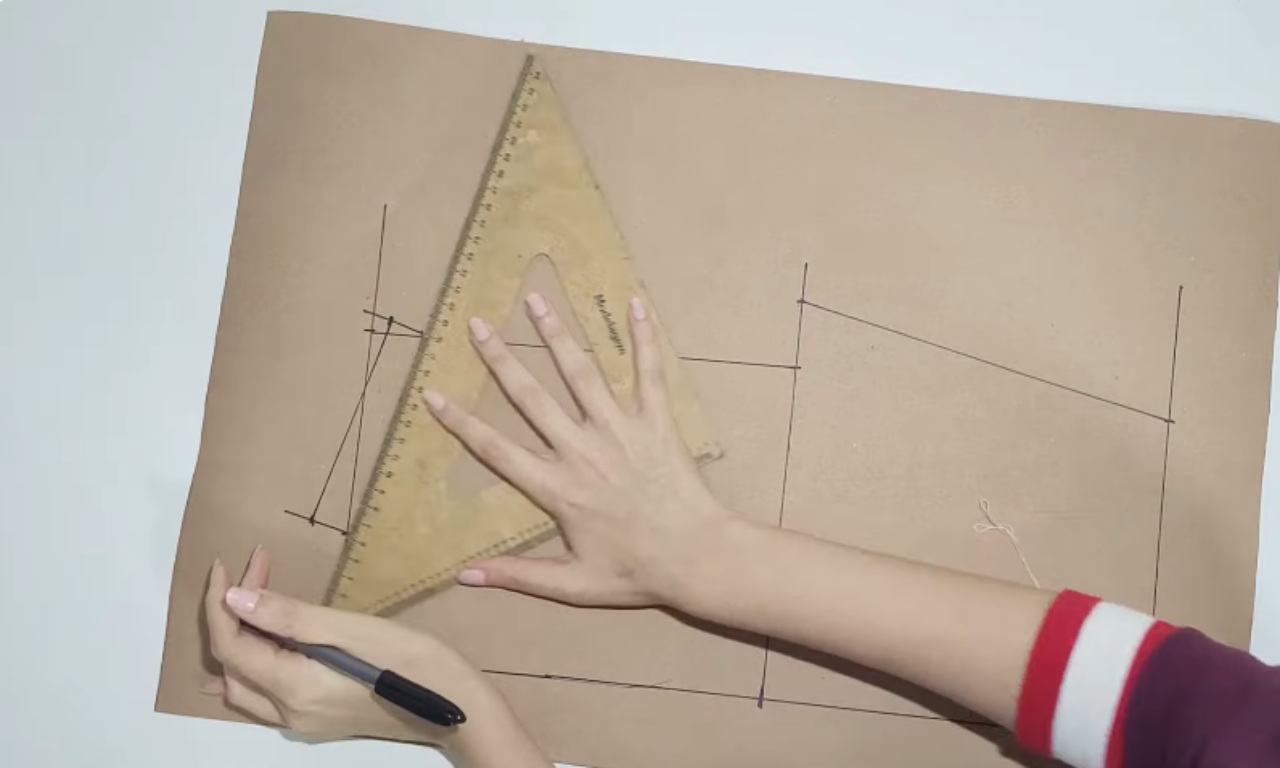 A hand using a triangular ruler and marker to draw straight lines on brown drafting paper for a clothing pattern.