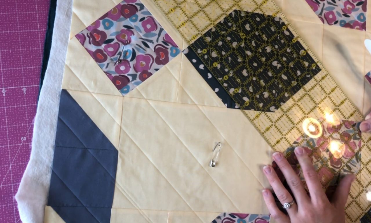 Close-up of hands aligning a quilting ruler on a patchwork quilt, with diagonal stitch lines visible on cream fabric squares and safety pins holding layers together.