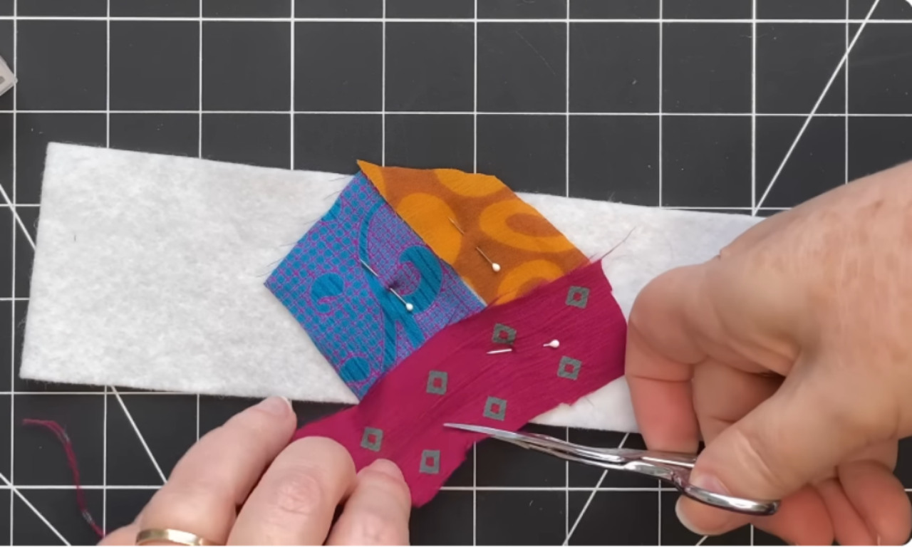The first step of making a modern crazy quilt: hands pinning and trimming colorful fabric scraps directly onto a white felt base to create a patchwork collage.