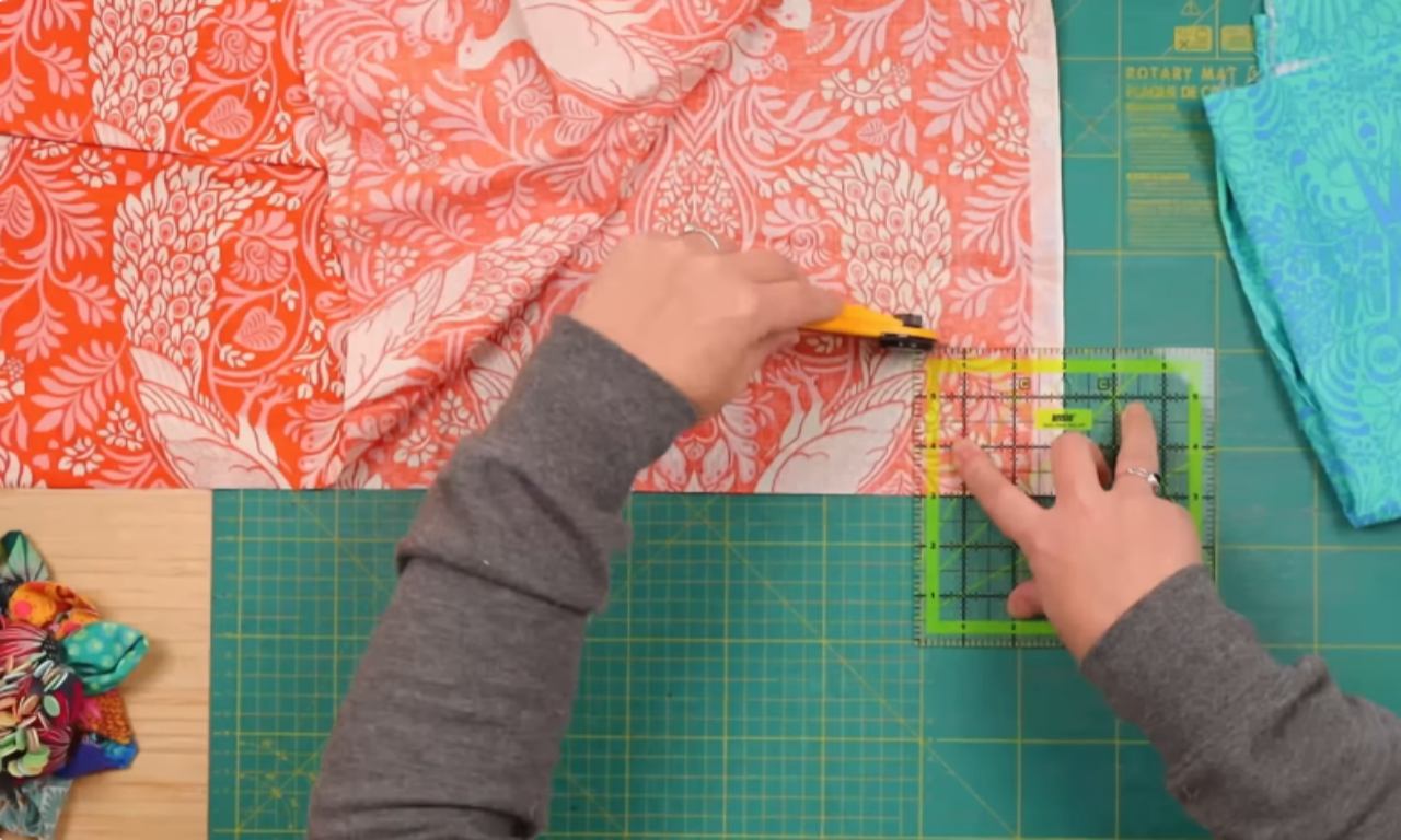 A person cuts a piece of coral and white patterned fabric using a rotary cutter and a square quilting ruler on a green cutting mat. Brightly colored fabric scraps and folded fabric are visible nearby.