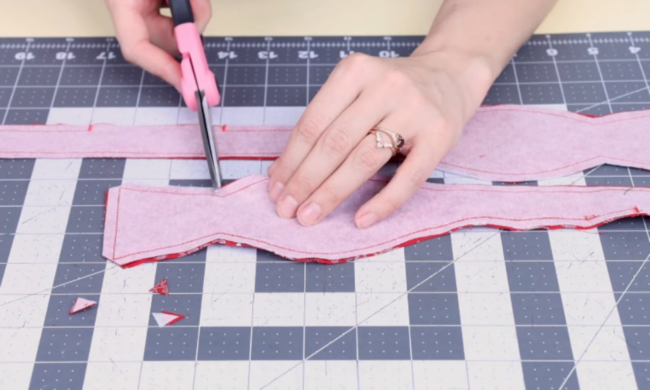 A person uses pink scissors to clip the seam allowance on a sewn piece of red bow tie fabric, which is laying on a grey cutting mat.