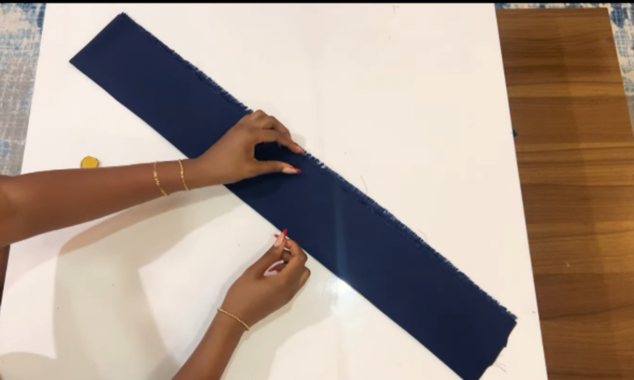 A person's hands folding a long, rectangular strip of dark blue fabric on a white cutting table, a step in sewing the tiered halter dress.